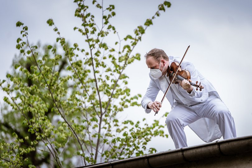 Ein Mann im weißen Anzug steht auf dem Dach eines Hauses und spielt auf seiner Geige. Grüne Baumkronen verschwimmen im Hintergrund. 
