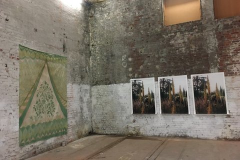 In einer Halle hängen in der Ecke eine Textilarbeit sowie drei nahezu identische Fotografien, auf denen Pflanzen zu sehen sind, nebeneinander an der unverputzten Ziegelsteinwand. 