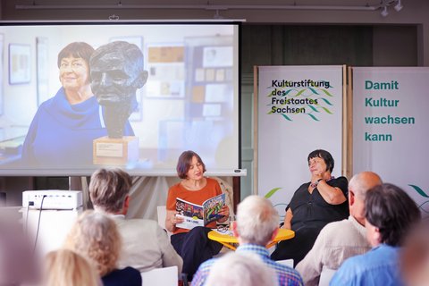 Franziska Gerstenberg liest vor Publikum aus einem Buchmagazin. Hinter ihr wird auf einer Leinwand ein Foto projiziert. Daneben ist das Logo der Kulturstiftung auf einem aufgestellten Banner zu sehen.