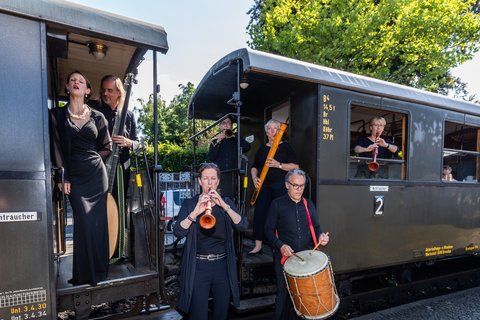 Eine Musikkapelle, ganz in schwarz gekleidet, steht vor und in einem historischen Eisenbahnwagon.