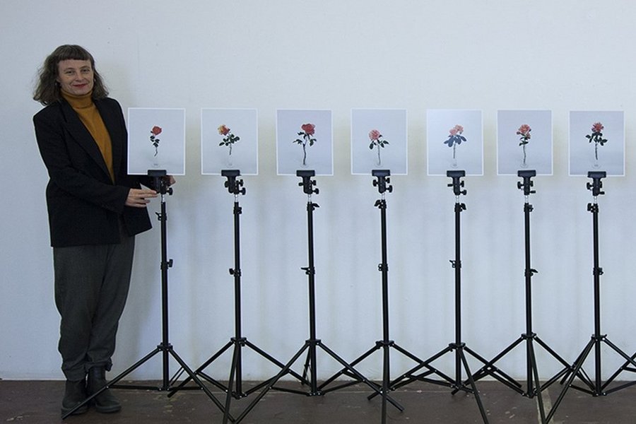 Die Künstlerin Antje Seeger steht vor einer weißen Wand neben in einer Reihe angeordneten Fotografien, die je an einem Stativ befestigt und auf denen Rosen abgebildet sind.