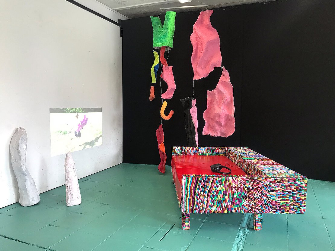 Kunstinstallation bestehend aus einem bunten Sofa und anderen farbigen Objekten sowie einer Projektion auf eine Wand.