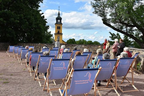 Eine Gruppe von älteren Menschen sitzt auf Liegestühlen, welche auf einer Schotterfläche aufgereiht wurden. Im Hintergrund ist ein gelber Kirchturm zu sehen.