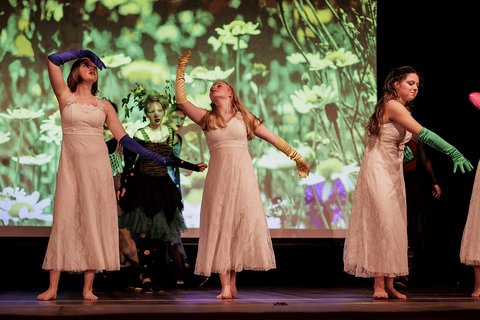 Tänzerinnen in weißen Kleidern führen ein Stück auf einer Bühne auf. Im Hintergrund wird auf eine Leinwand eine Blumenwiese projiziert.