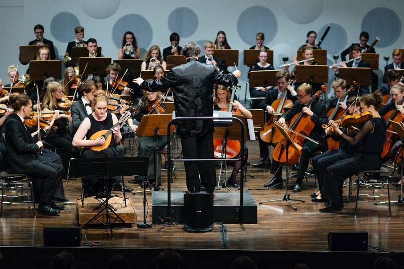 Ein in schwarz gekleidetes Orchester mit Dirigenten spielt auf einer Bühne vor Publikum.