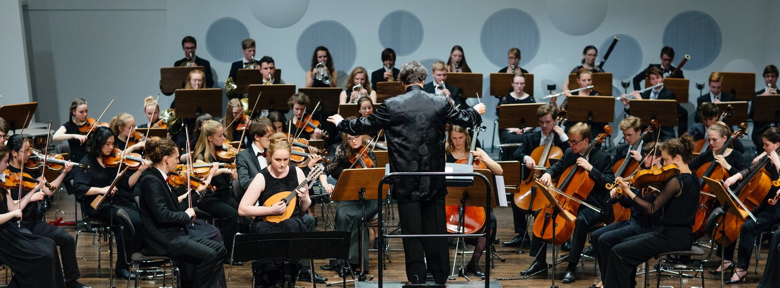 Ein in schwarz gekleidetes Orchester mit Dirigenten spielt auf einer Bühne vor Publikum.