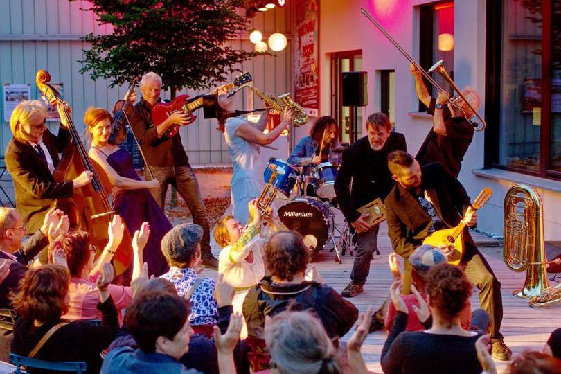 Eine Band mit 8 Musizierenden spielt vor applaudierendem Publikum in einer fröhlich ausgelassenen Stimmung.