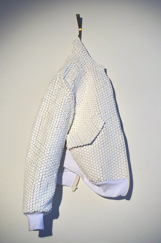 Eine weiße Bomberjacke hängt an einem Haken. Statt Stoff sind weiße Tabletten sorgfältig aneinandergenäht. 