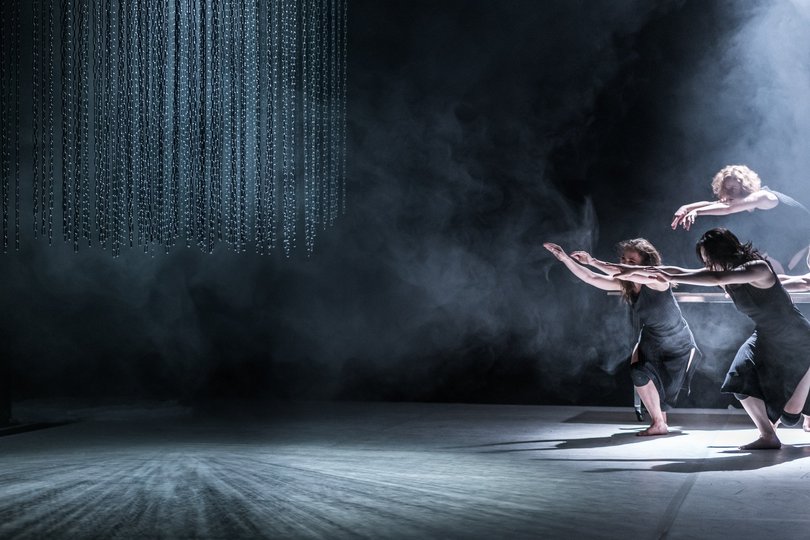 Vier Tänzerinnen im Lichtkegel auf einer verdunkelten Bühne, sie tragen schwarze Sachen