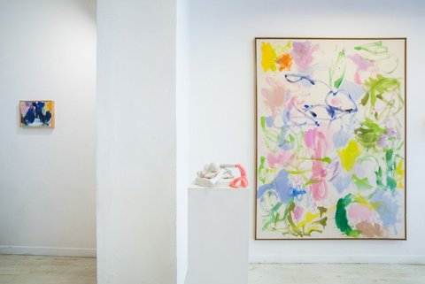 In einem Ausstellungsraum hängt ein abstraktes, farbiges Gemälde an der Wand. Davor steht eine weiße Stele mit einer Plastik darauf.