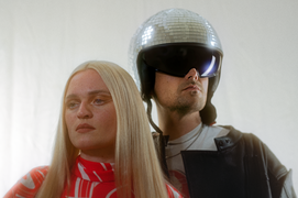 Eine Frau mit glatten blonden Haaren und ernstem Gesichtsausdruck steht neben einem Mann mit silbernem Discohelm und einer schwarzen Sonnenbrille.