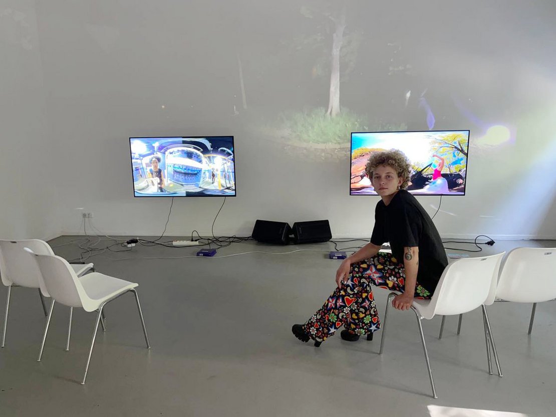 Eine Frau sitzt auf einem Stuhl und blickt rückwärts in die Kamera, hinter ihr hängen zwei Bildschirme an der Wand des leeren Raumes.