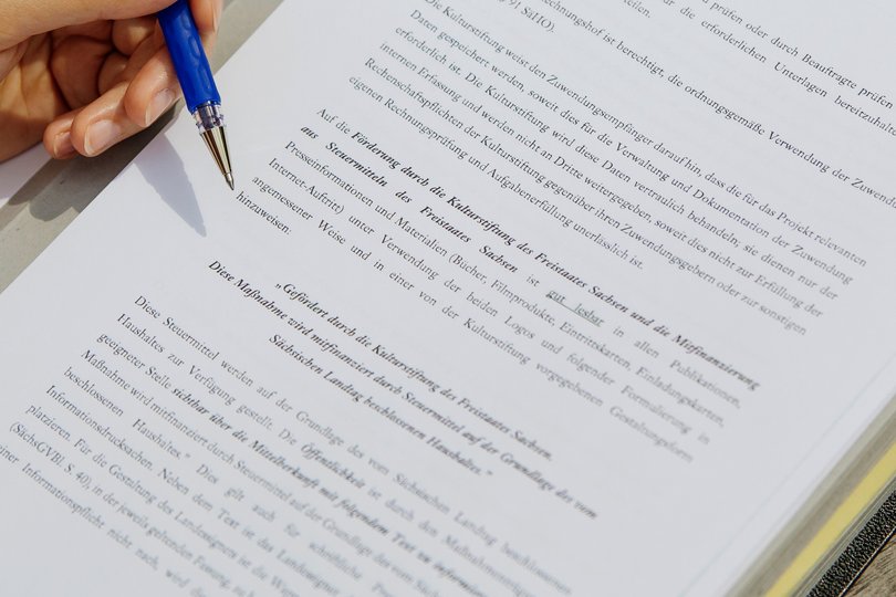Eine Hand mit Stift schwebt über einem digital beschrifteten Blatt Papier. 